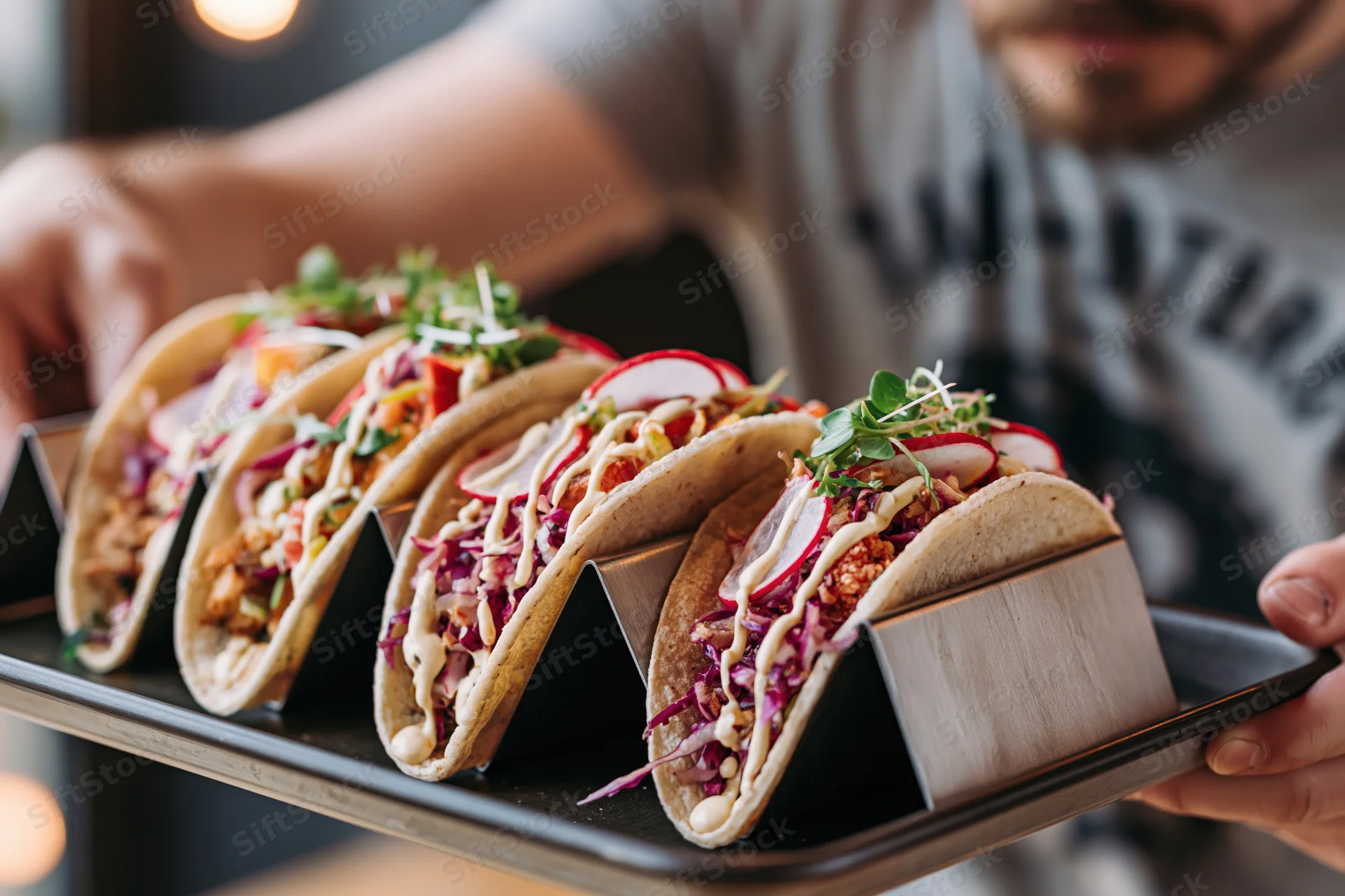 Man holding tray with four gourmet tacos in metal holders, each filled with meat, cabbage, radish, and sauce, arranged in a row