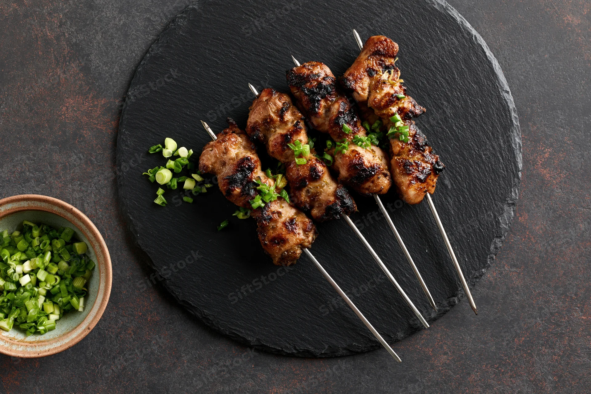 Grilled chicken skewers on metal rods arranged on a round slate plate with chopped green onions in a ceramic bowl beside it