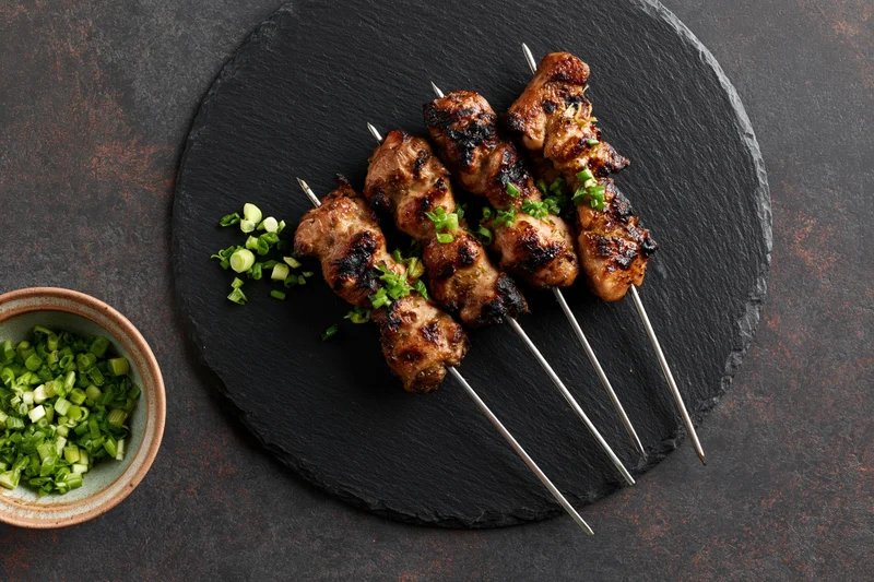 Grilled chicken skewers on metal rods arranged on a round slate plate with chopped green onions in a ceramic bowl beside it