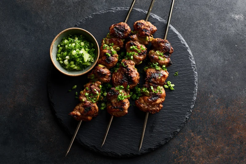 Grilled chicken skewers on metal rods arranged on a round slate plate with chopped green onions on the side