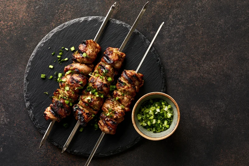 Grilled chicken skewers on metal rods arranged on a slate platter with chopped green onions on the side