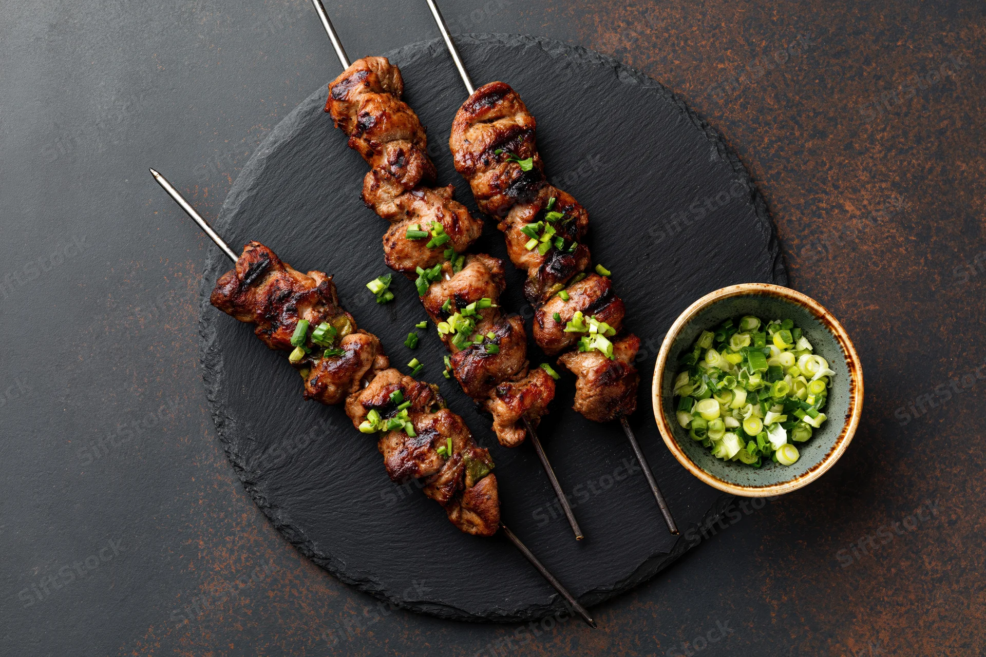 Grilled chicken skewers on metal rods arranged on a slate platter with chopped green onions in a ceramic bowl