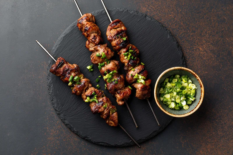 Grilled chicken skewers on metal rods arranged on a slate platter with chopped green onions in a ceramic bowl