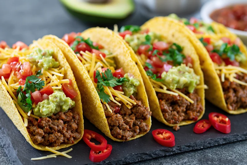 Four hard-shell tacos filled with seasoned ground beef, cheese, guacamole, tomatoes, and cilantro arranged on a slate plate with sliced chili peppers nearby