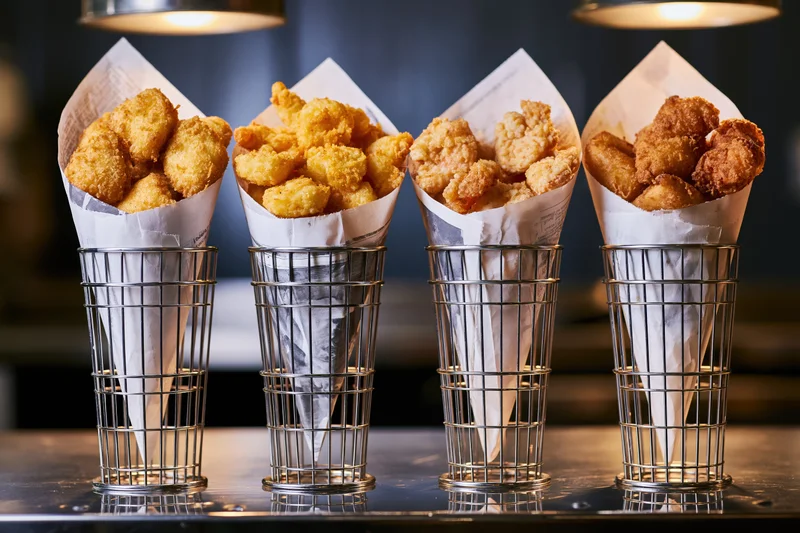 Four metal cone holders each filled with fried chicken in paper wrappers on a reflective restaurant counter under warm overhead lights