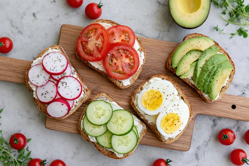 Five open-faced toasts with avocado, tomato, cucumber, radish and egg on a wooden board with cherry tomatoes and herbs around