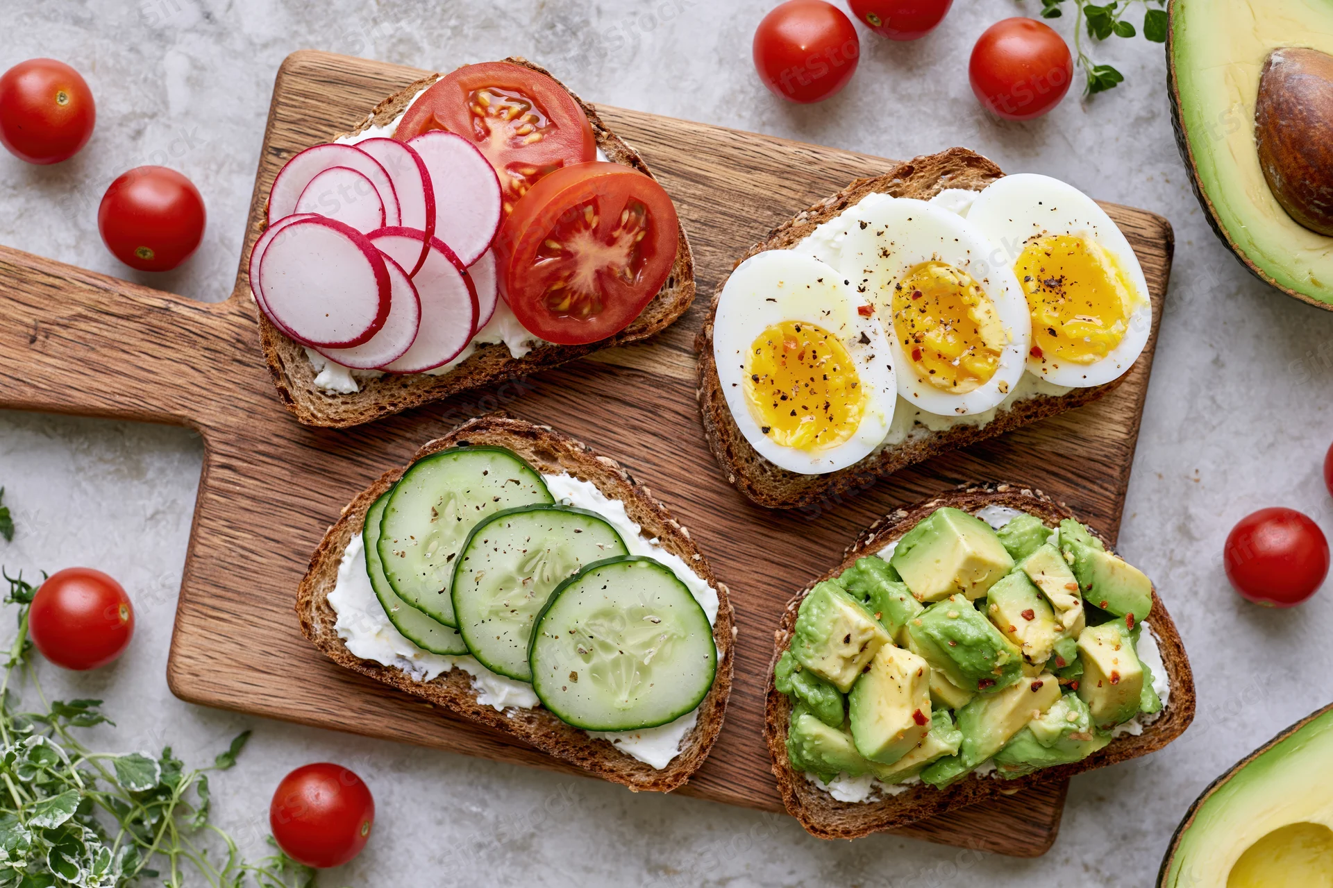 Four open-faced toasts with avocado, egg, cucumber, radish and tomato on a wooden cutting board