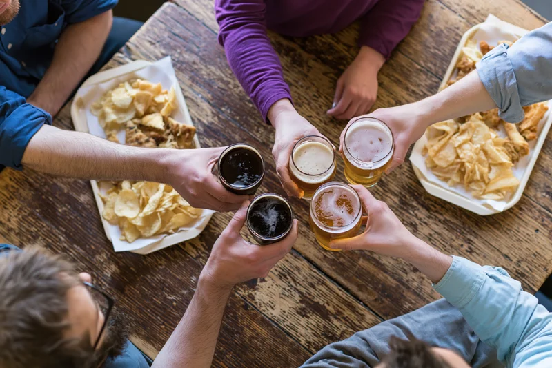 Friends raising beer glasses in toast around a wooden pub table with snacks
