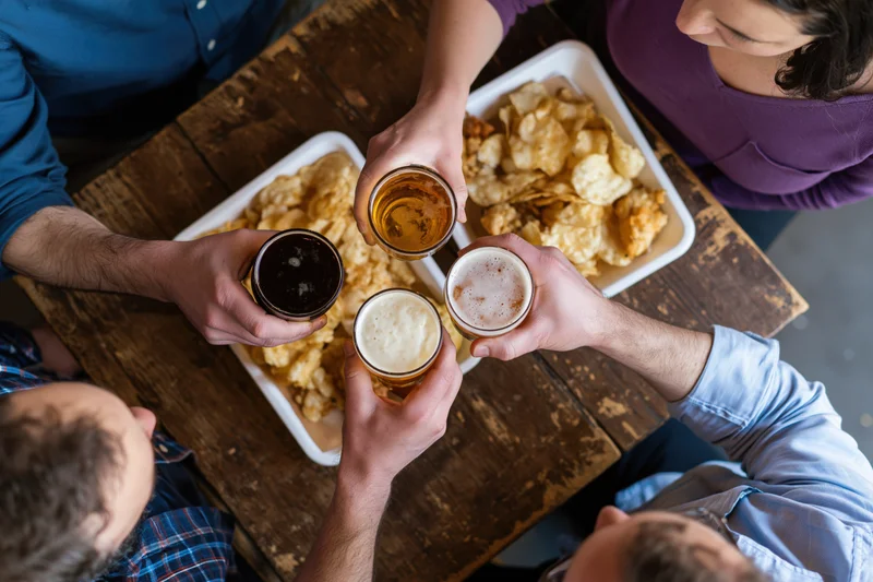 Four people raising beer glasses in a toast around a wooden pub table with snacks