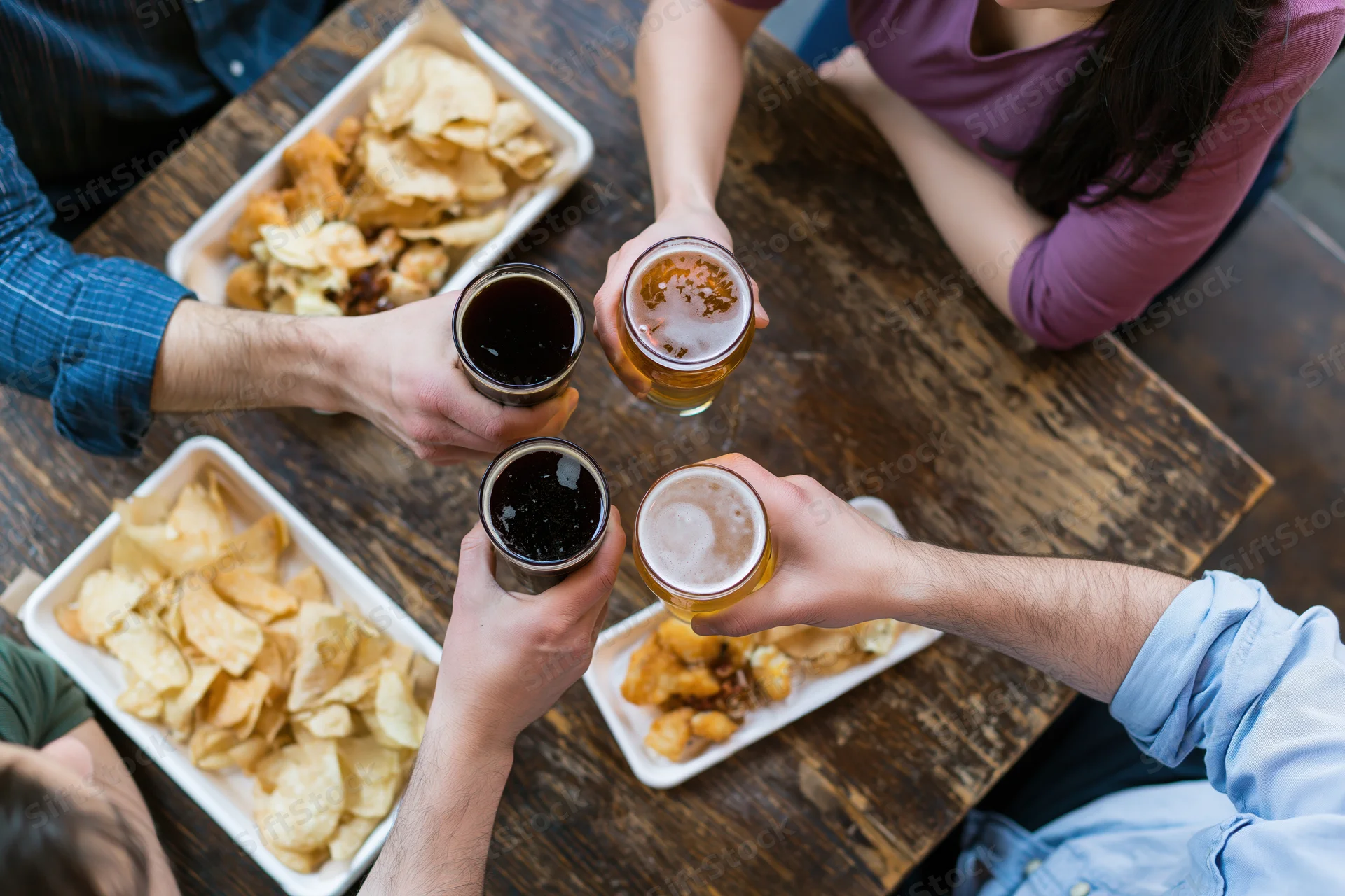 Four people toasting with beer glasses over a wooden pub table surrounded by trays of chips and fried snacks