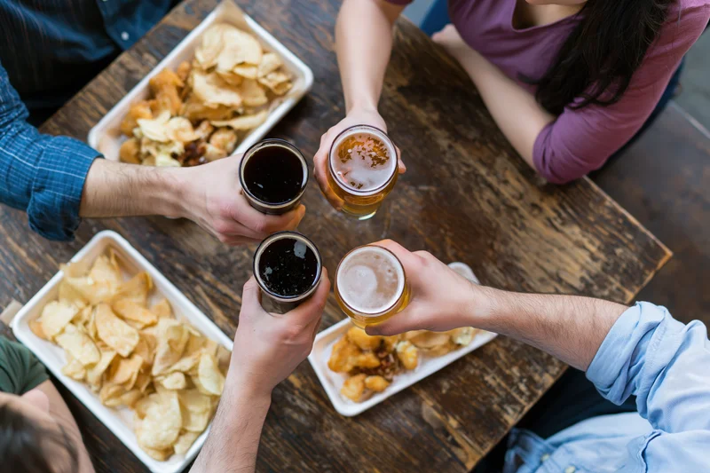 Four people toasting with beer glasses over a wooden pub table surrounded by trays of chips and fried snacks