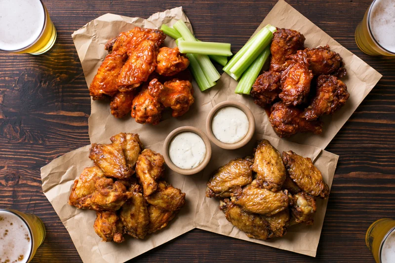 Four portions of chicken wings in different sauces arranged on parchment paper with celery sticks and ranch dip on a wooden table, surrounded by glasses of beer