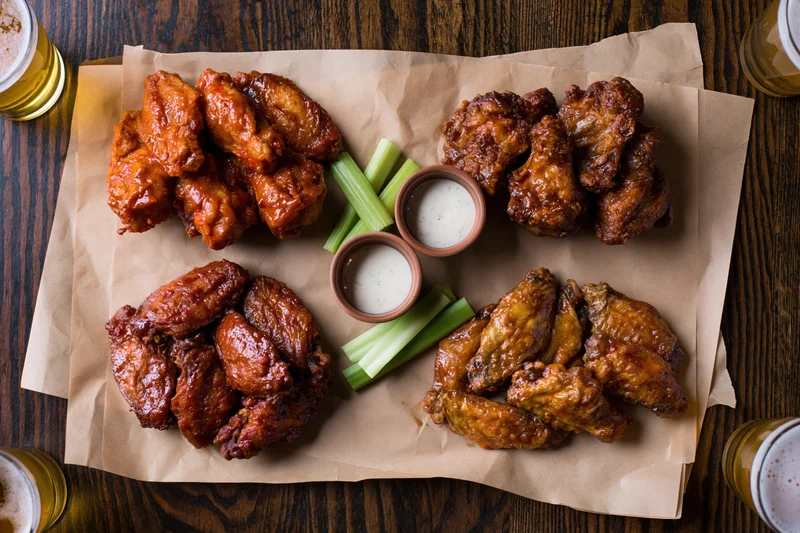 Four portions of chicken wings in different sauces arranged on parchment paper with celery sticks and ranch dressing on a wooden table, surrounded by glasses of beer