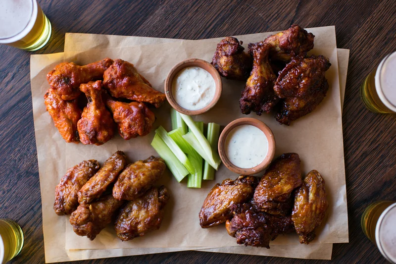 Four portions of chicken wings in different sauces arranged on parchment paper with celery sticks and ranch dip on a wooden table, surrounded by glasses of beer