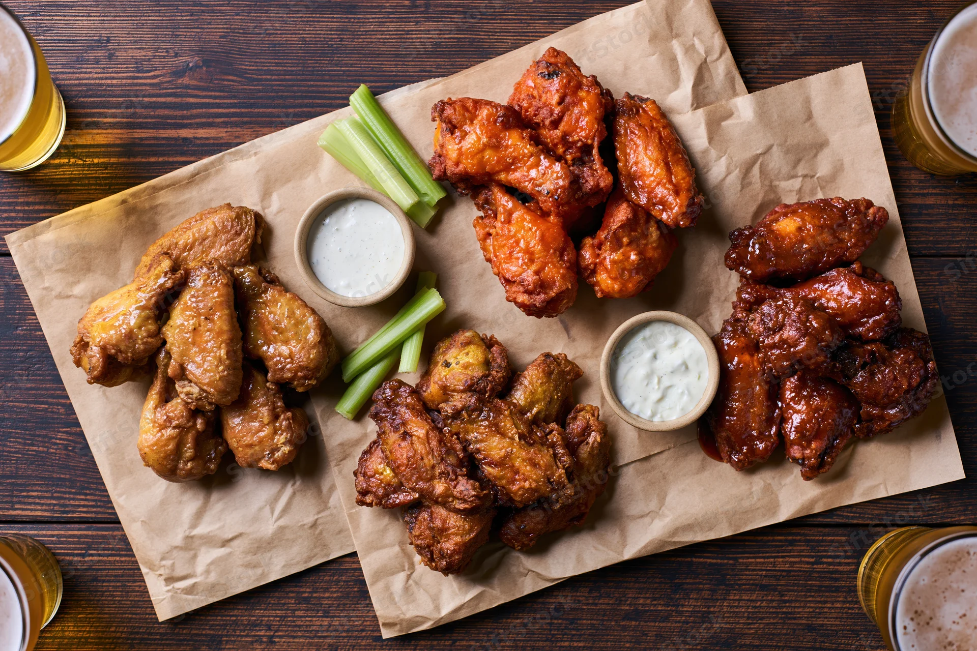 Four portions of chicken wings in different sauces arranged on brown paper with celery sticks and ranch dip on a wooden table, surrounded by glasses of beer