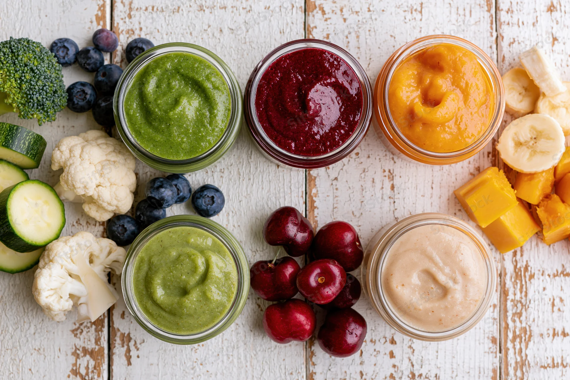 Four glass jars of colorful baby food puree arranged with fresh fruits and vegetables on a rustic wooden table
