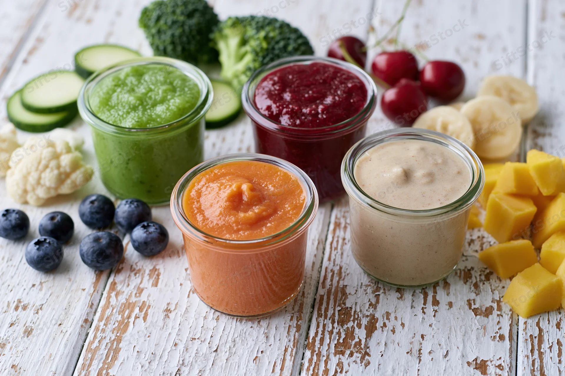Four glass jars of colorful baby food purees arranged on a rustic white wooden table with fresh vegetables and fruits around