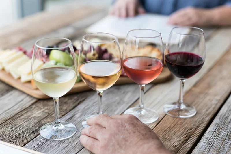 Four wine glasses in a row holding white, rosé, and red wines on a wooden table with a cheese and charcuterie board in the background