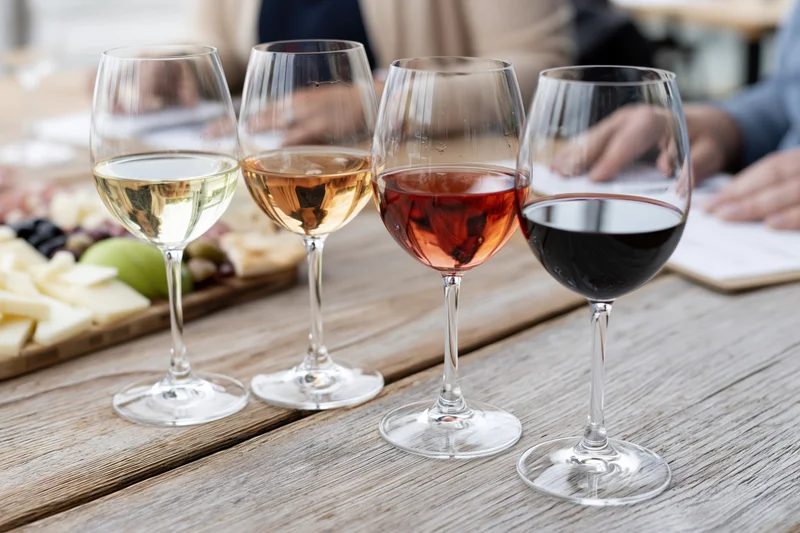 Four wine glasses in a row holding white, rosé, and red wines on a wooden table with cheese and charcuterie board in background