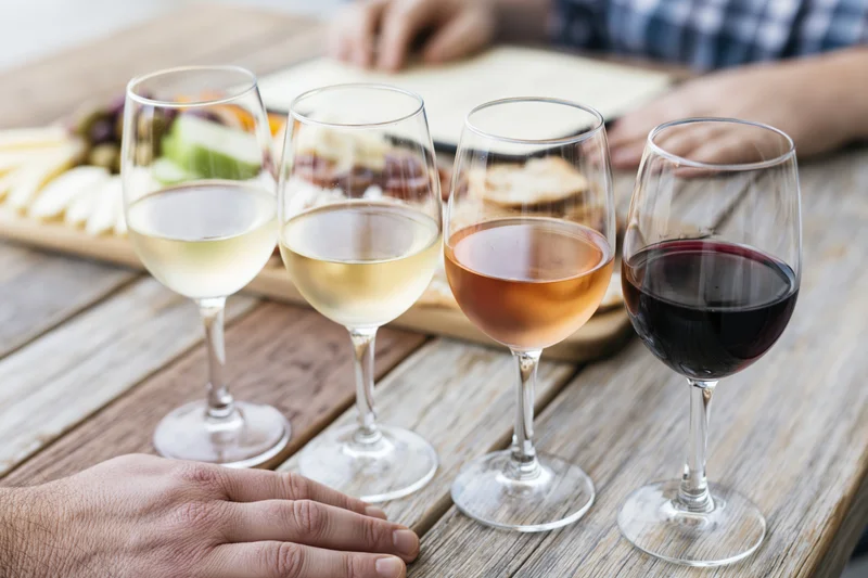 Four wine glasses with white, rosé and red wine arranged in a row on a wooden table with cheese and charcuterie platter in background