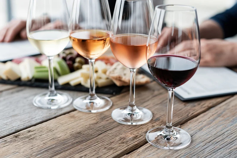 Four wine glasses with white, rosé and red wine arranged in a row on a wooden table beside a charcuterie platter