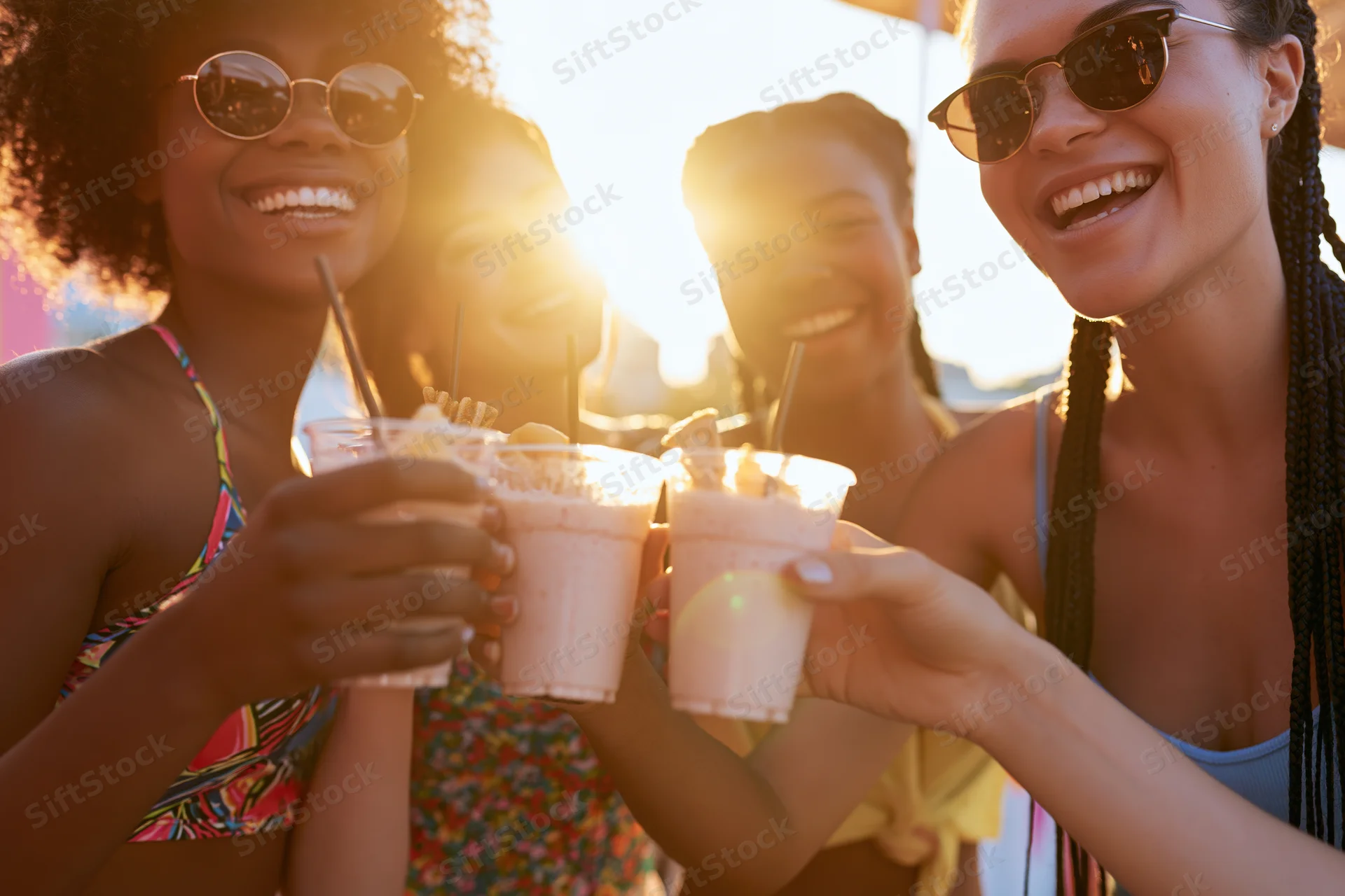 Four young women smiling and clinking plastic cups at sunset on the beach
