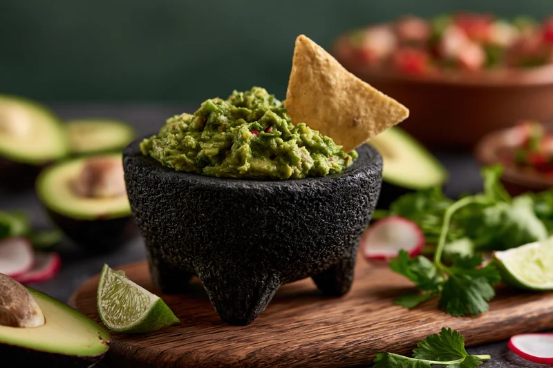 Fresh guacamole in a black stone molcajete with a tortilla chip on a wooden board surrounded by ingredients