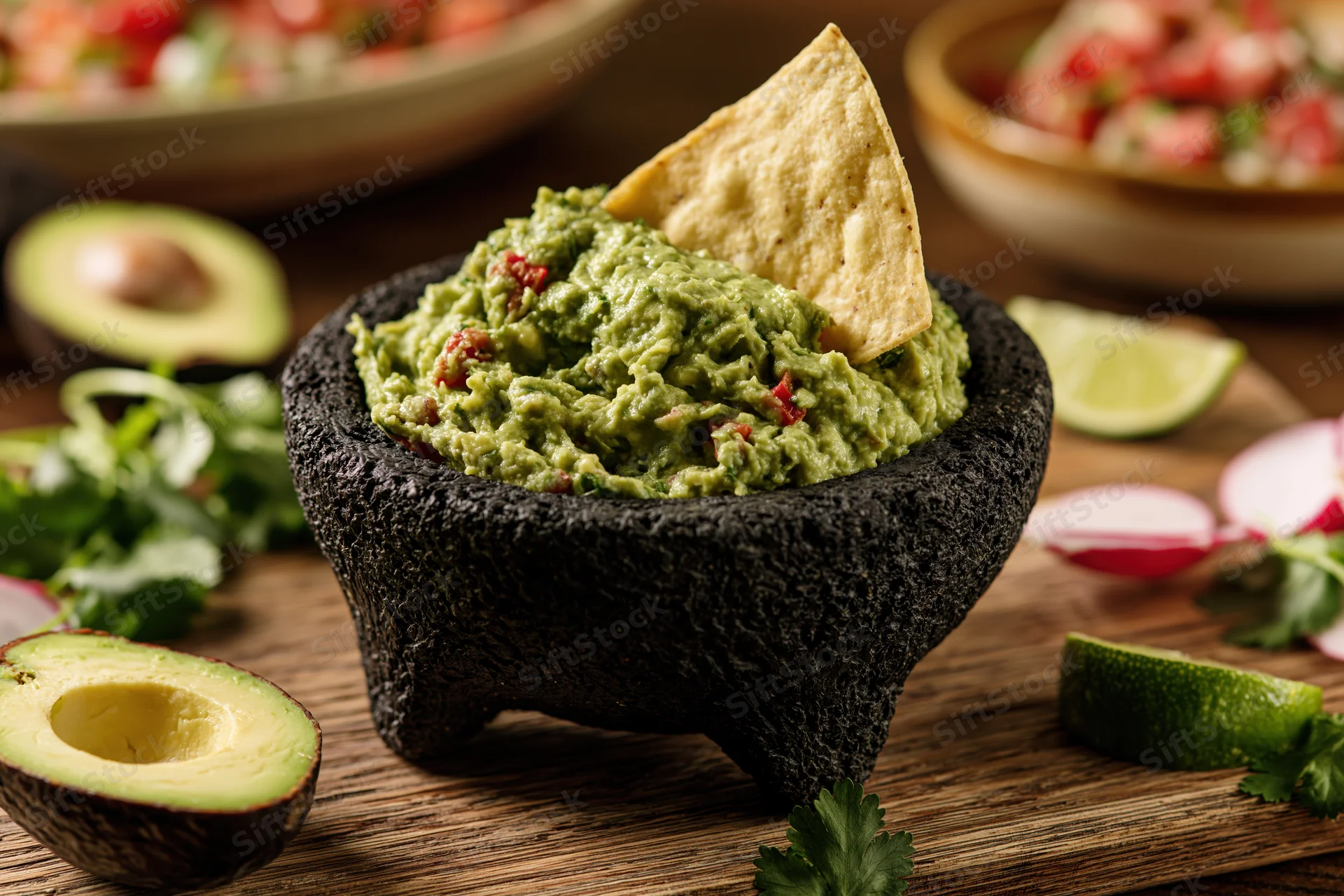 Fresh guacamole in a carved black stone molcajete with a tortilla chip on a wooden table surrounded by ingredients