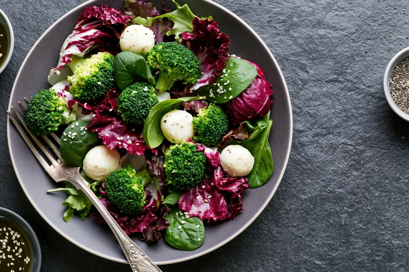 Fresh salad with broccoli and mozzarella balls on a gray plate with fork and sesame seeds on dark stone surface
