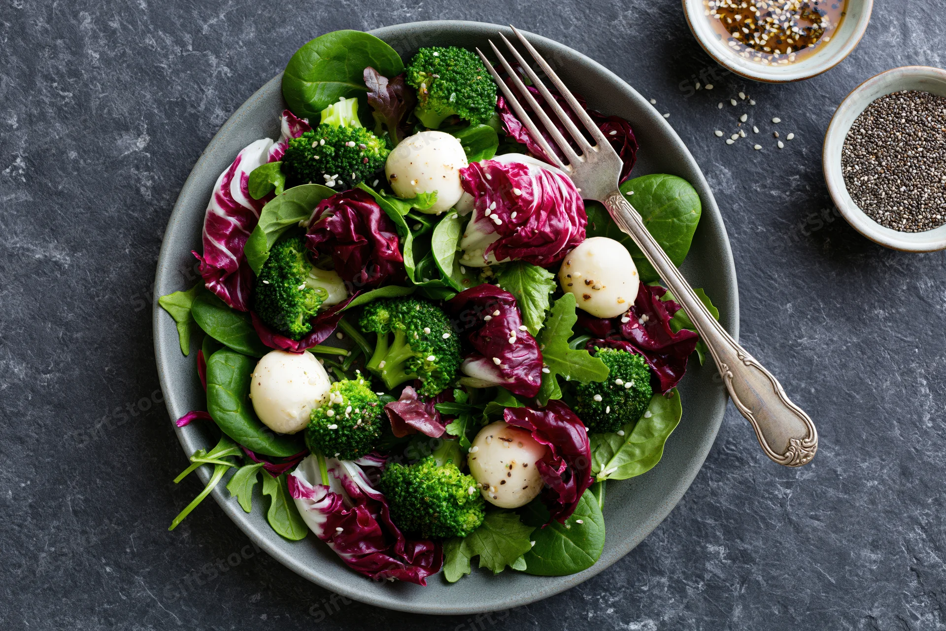 Salad with broccoli, mozzarella balls and radicchio on a gray stone surface with a fork and small bowls of seeds and dressing nearby