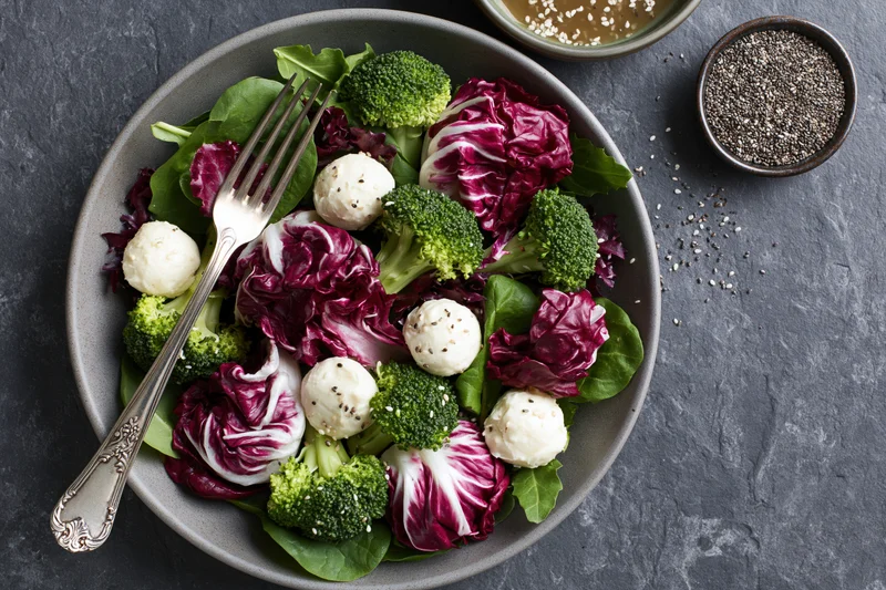 Salad bowl with mozzarella balls, broccoli, and radicchio on gray stone surface with fork and small bowls of seeds and dressing beside it