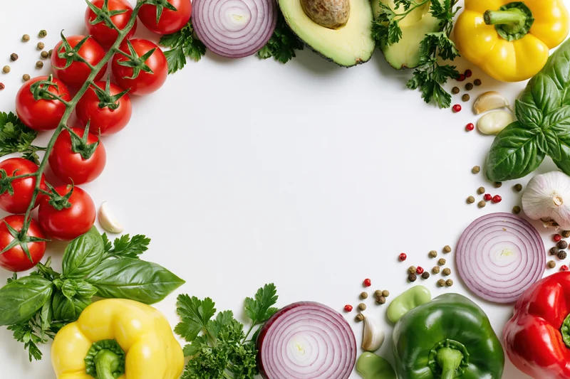 Fresh vegetables and herbs arranged in a loose circular border on white surface with copyspace