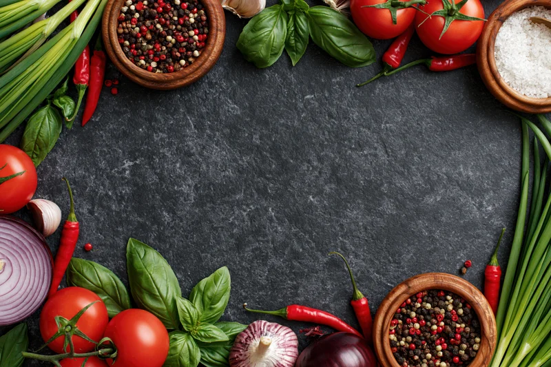 Fresh vegetables and spices arranged in a border frame around a dark surface with copyspace in center