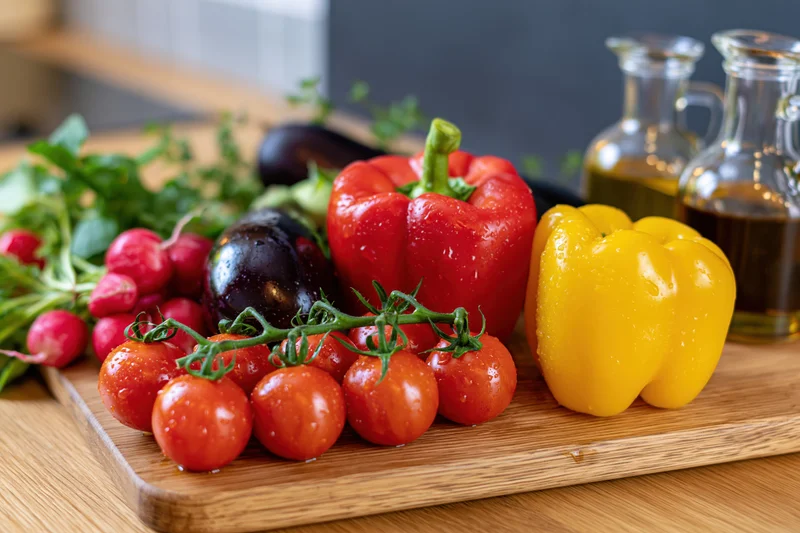 Fresh vegetables including tomatoes, bell peppers, radishes and eggplant arranged on a wooden cutting board in a kitchen setting