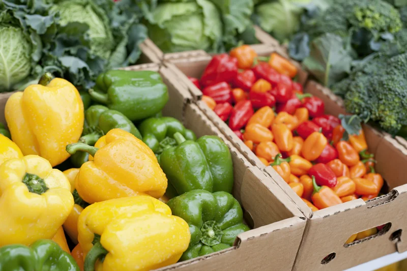 Fresh bell peppers and leafy vegetables in open cardboard boxes at a market display
