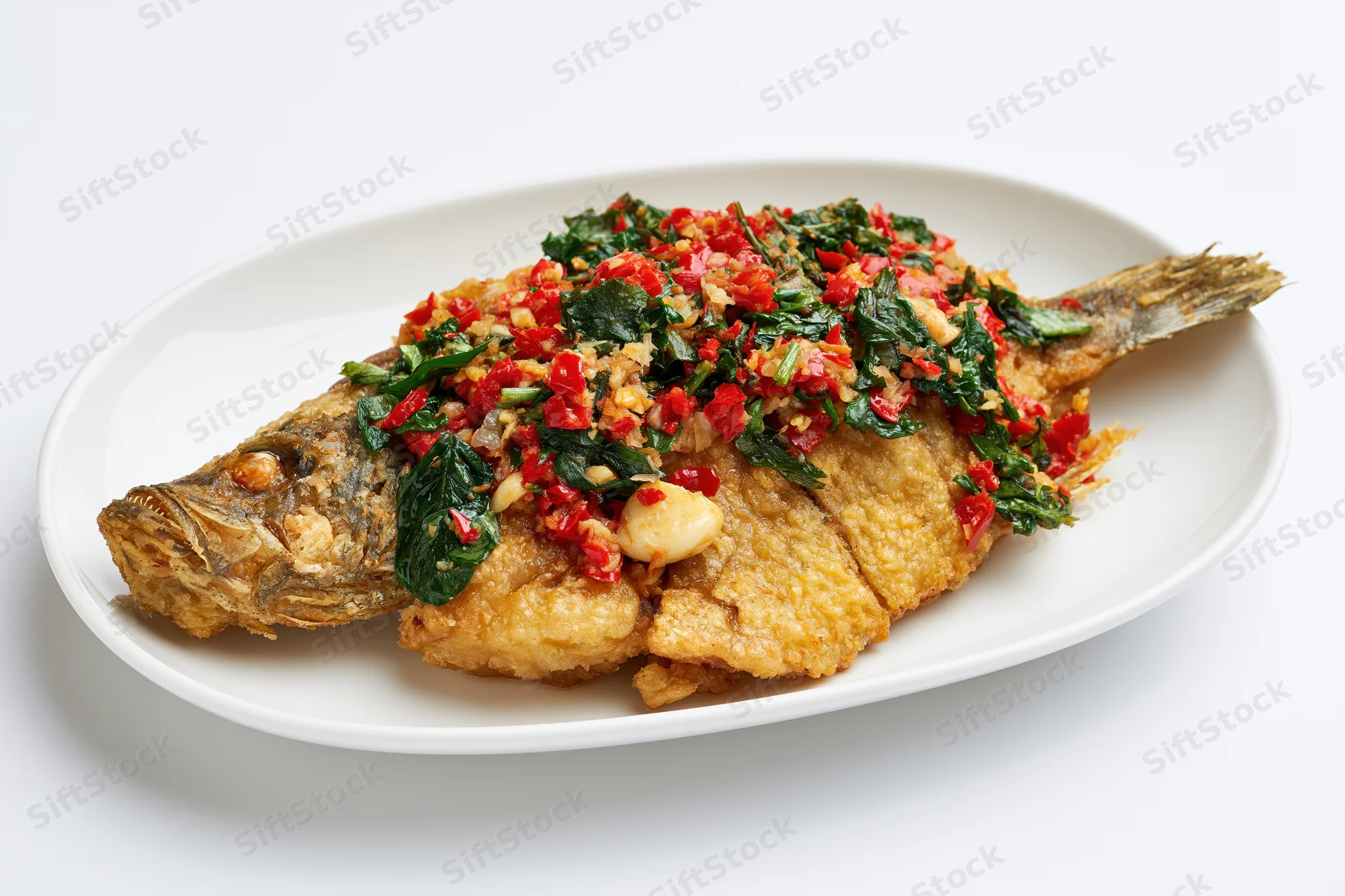 Whole fried fish topped with stir-fried thai basil and chili on a white plate