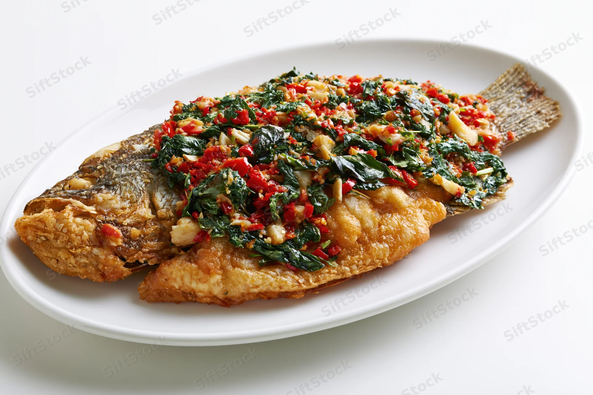 Whole fried fish topped with stir-fried thai basil and chili on a white plate