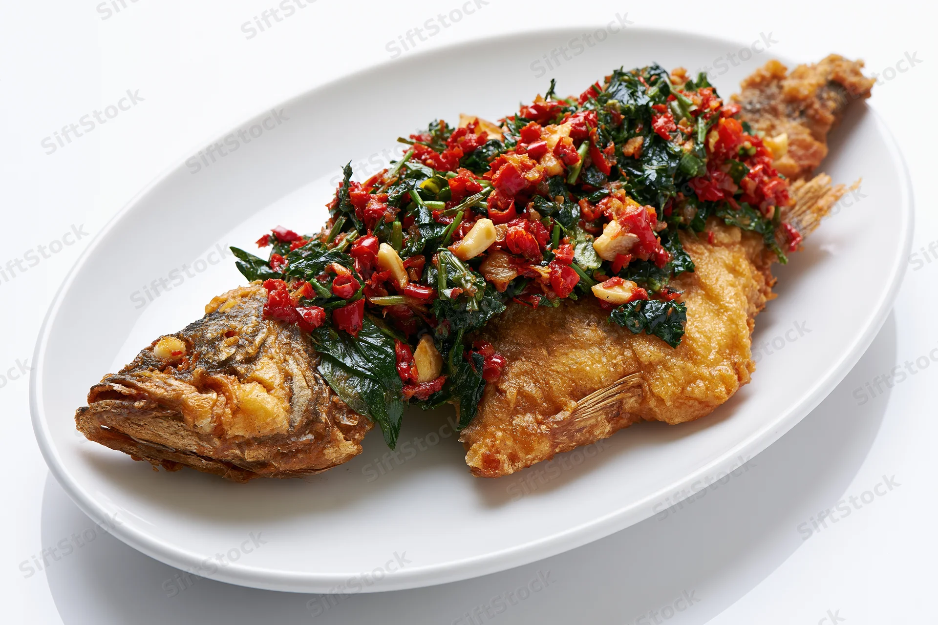 Whole fried fish topped with stir-fried thai basil and red chili on a white oval plate