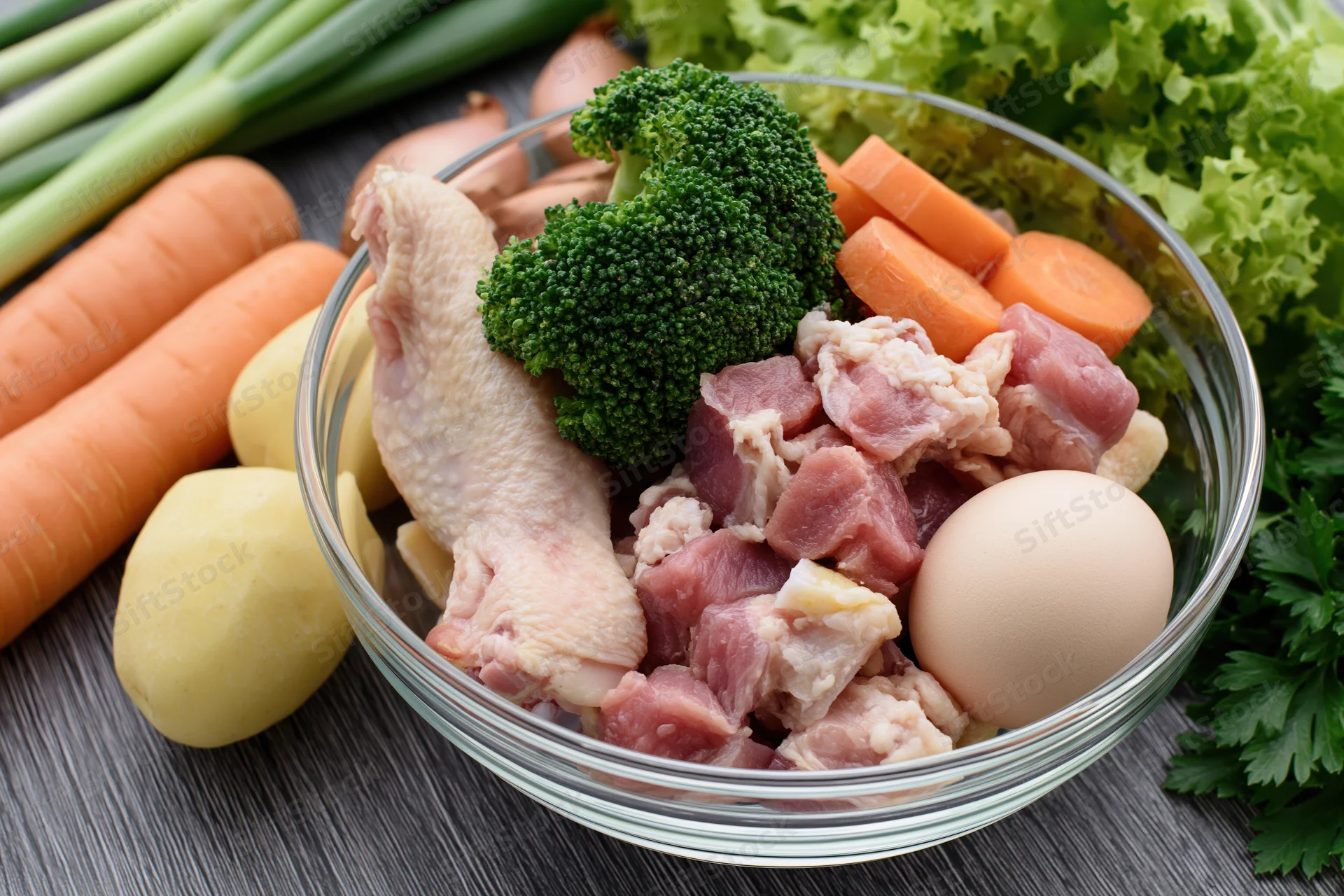 Glass bowl with raw chicken, diced meat, broccoli, carrot, egg and potato on wooden surface surrounded by fresh vegetables