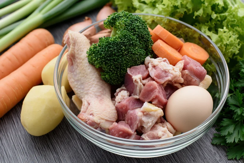 Glass bowl with raw chicken, diced meat, broccoli, carrot, egg and potato on wooden surface surrounded by fresh vegetables