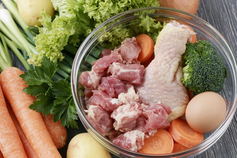 Glass bowl with raw chicken, diced meat, broccoli, carrots, egg and potatoes on wooden surface surrounded by fresh vegetables
