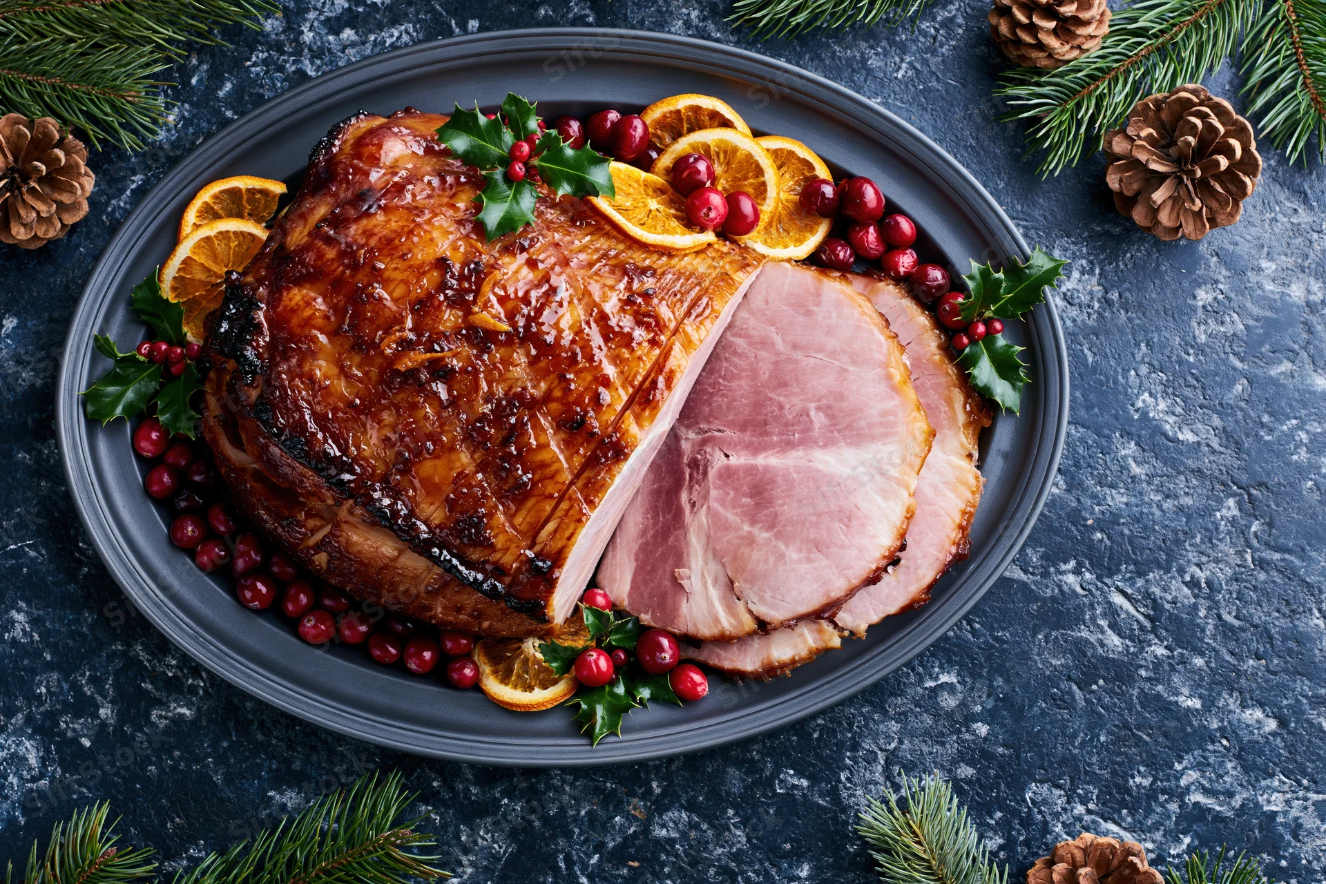 Glazed ham with a slice cut resting on a dark platter surrounded by holly, cranberries, orange slices, and pinecones for christmas dinner