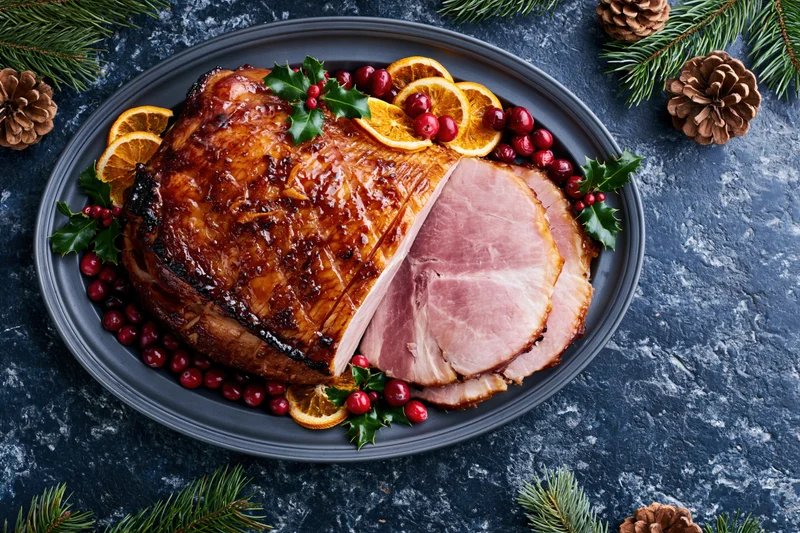 Glazed ham with a slice cut resting on a dark platter surrounded by holly, cranberries, orange slices, and pinecones for christmas dinner