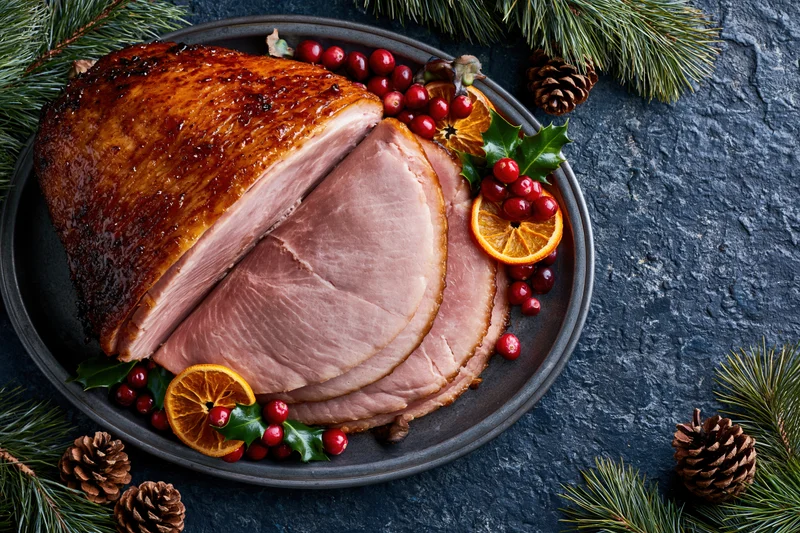 Glazed ham with cranberries and orange slices on a dark stone plate surrounded by pine branches and pinecones