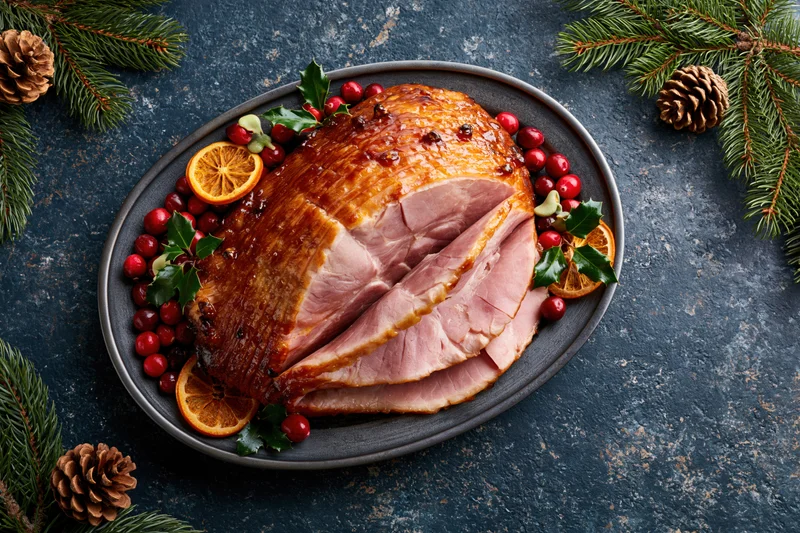 Glazed ham with cranberries and orange slices on a dark plate surrounded by pine branches and pinecones
