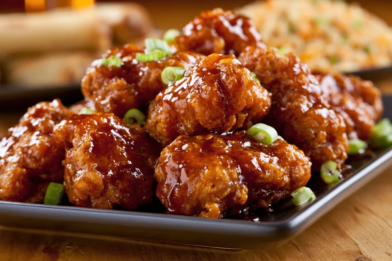 Glazed orange chicken pieces piled on a dark square plate with green onion garnish on wooden table