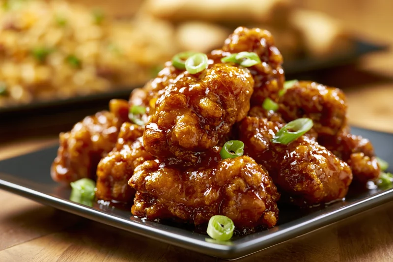 Glazed orange chicken pieces piled on a dark square plate with green onion garnish on a wooden table