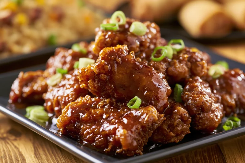 Glazed orange chicken pieces piled on a dark square plate with green onion garnish on a wooden table