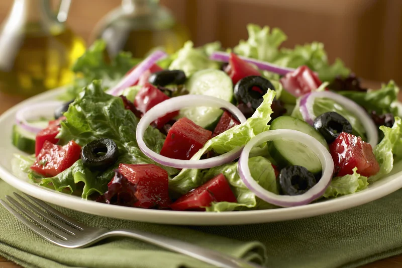 Greek-style salad with lettuce, tomato, cucumber, olives and red onion on a white plate with fork and napkin
