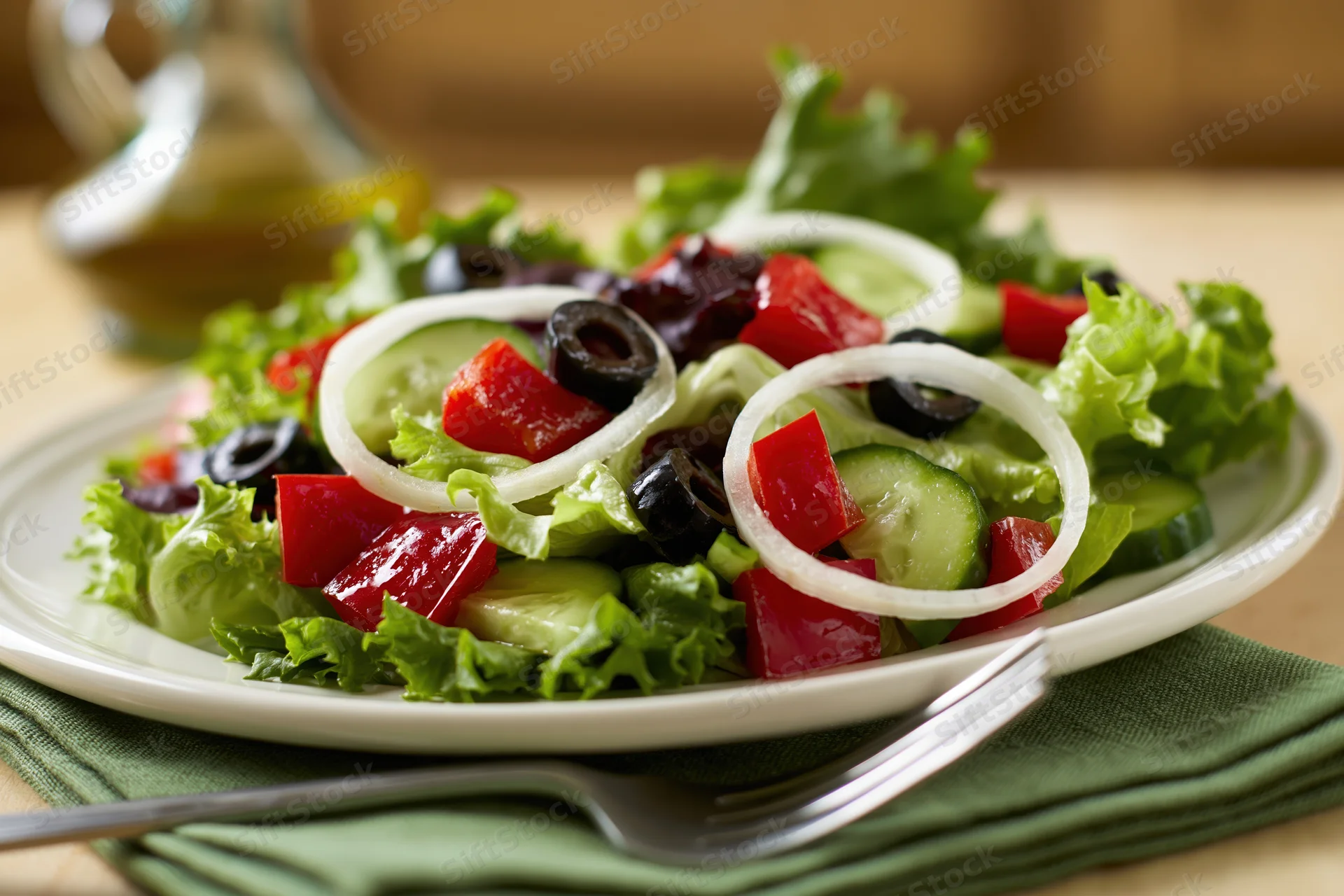 Greek-style salad with lettuce cucumber tomatoes olives and onions on a white plate with fork and green napkin on table